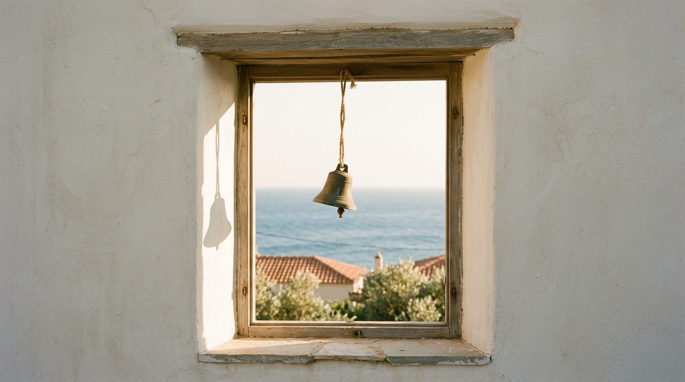A small weathered bronze bell hanging on a cord in a bright open window, its shadow cast on the stucco wall
