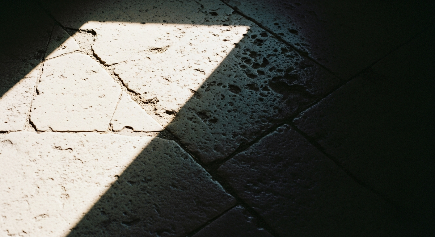 A stone floor with a sharp diagonal shadow line dividing bright from dark, a small pile of dust scattered in the light zone